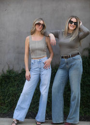 Two women posing outdoors against a concrete wall with ivy, wearing sunglasses, neutral tops and high-waisted wide-leg jeans — casual urban street-style look.