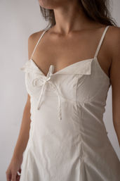 Close-up of a white spaghetti-strap summer dress with a sweetheart neckline and tied bow at the center bust, fitted seam details and flowing skirt on a neutral studio background.