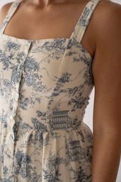 Close-up of a person's torso wearing a cream button-front summer sundress with blue toile pastoral print, square neckline, wide straps and gathered waist.