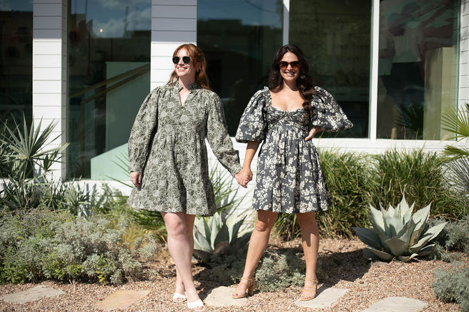 Two smiling women in sunglasses holding hands outside a white storefront, wearing short floral dresses with puffed sleeves and standing on a sunny gravel garden with agave and succulents.