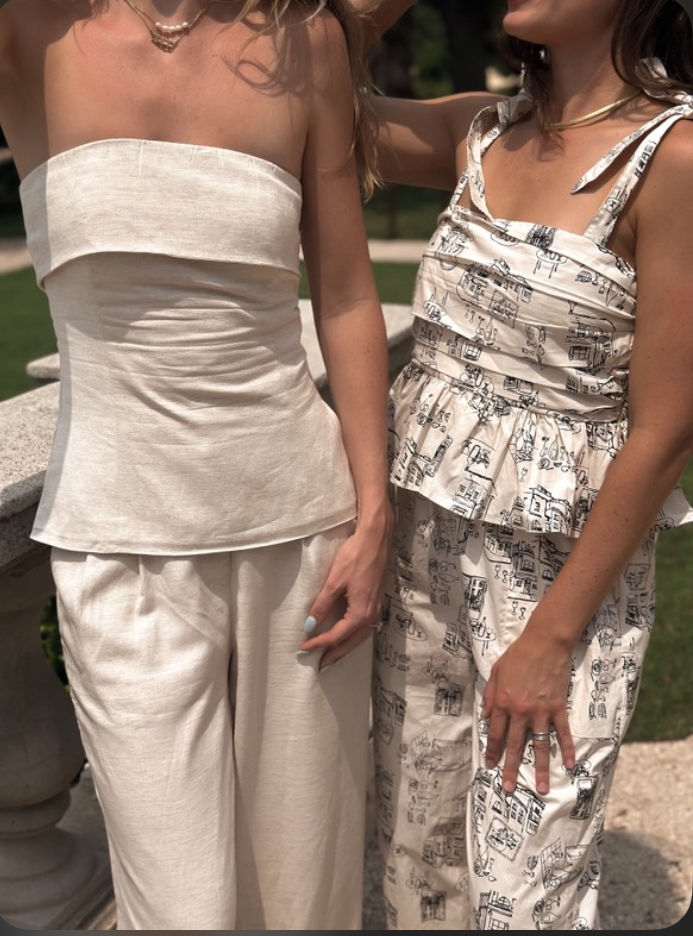 Two women on a sunny garden terrace wearing neutral summer outfits — strapless linen top with wide-leg pants and a printed tie-shoulder peplum matching set.
