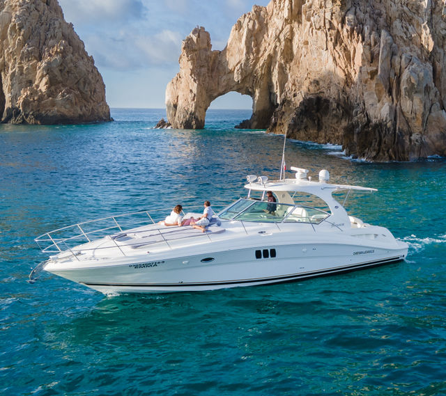 White luxury yacht cruising turquoise sea past a dramatic coastal rock arch, passengers lounging on the sun-soaked deck beneath a bright blue sky