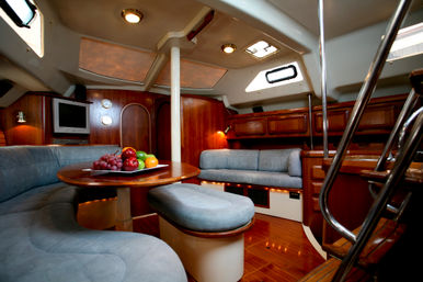 Cozy luxury yacht interior with curved gray upholstered seating around a round wooden table topped with a fruit platter, warm wood-paneled salon, porthole windows, skylights and stainless-steel stairs.