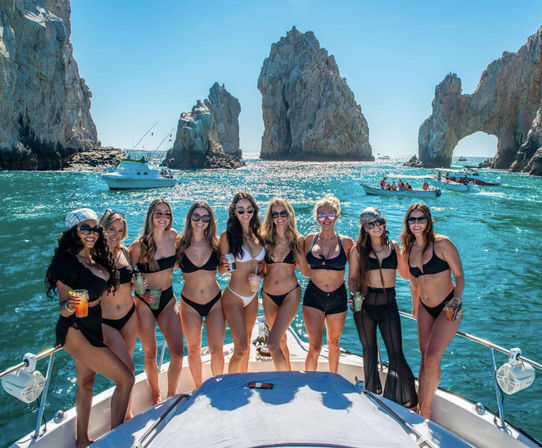 Ten bikini-clad women enjoying a boat party with drinks in front of El Arco rock formation at Lands End, Cabo San Lucas, Mexico, sunny turquoise sea.