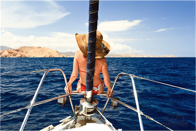 Sun-hatted traveler perched on the bow of a sailboat, facing deep-blue ocean and sunlit rocky coastline under a clear sky.