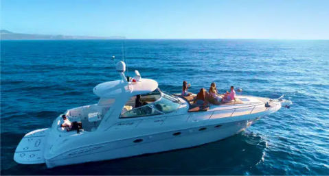 Group of people sunbathing and socializing on a white luxury motor yacht cruising on clear blue sea under a bright sunny sky with a distant coastline on the horizon