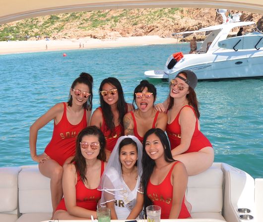 Bachelorette and six friends in matching red swimsuits posing on a yacht with turquoise water and a sandy beach cove in the background.