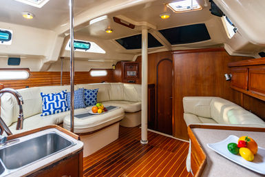 Cozy sailboat cabin interior with cream leather L-shaped seating, teak wood floors and paneling, compact galley sink, blue patterned pillows and bowls of fruit beneath skylights.