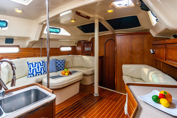 Cozy sailboat cabin interior with cream leather L-shaped seating, teak wood floors and paneling, compact galley sink, blue patterned pillows and bowls of fruit beneath skylights.