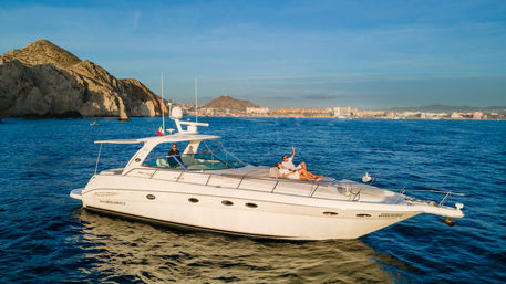 Sunlit white motor yacht on deep blue sea near rocky coastal cliffs, couple relaxing on the bow and a captain at the helm with a distant seaside skyline and hills under a clear blue sky — leisure yacht vacation scene.