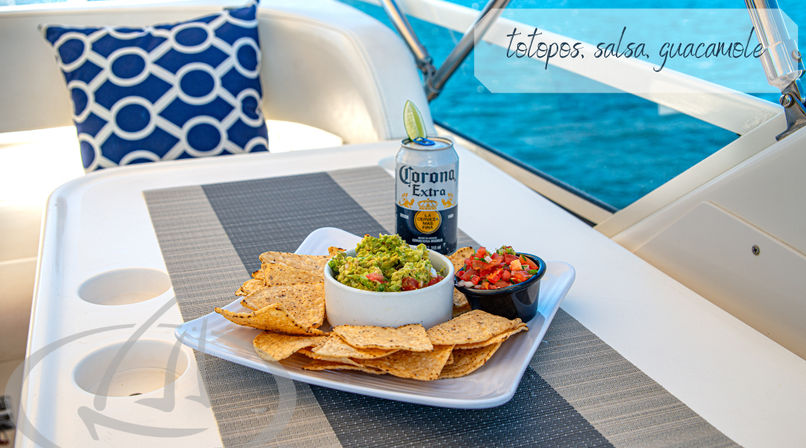 Plate of tortilla chips with bowls of guacamole and salsa beside a Corona Extra can with lime on a sunny boat deck, gray placemat and blue ocean in the background