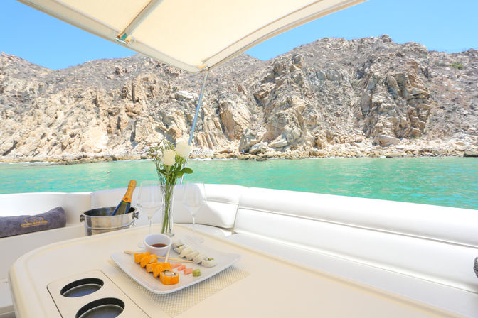 Luxury yacht deck with sushi platter, chilled champagne in an ice bucket, white roses on the table overlooking turquoise water and a rocky coastal cove under a clear blue sky