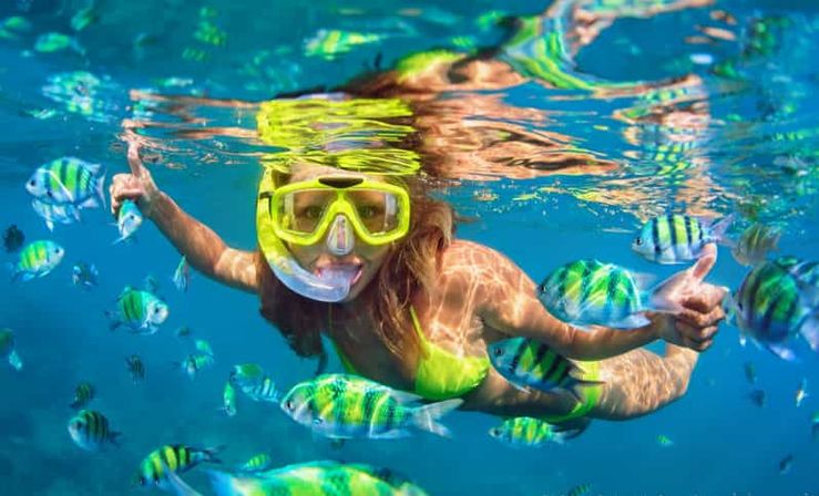 Snorkeler in neon yellow mask and green swimsuit giving a thumbs-up while swimming among colorful striped tropical fish in clear turquoise ocean water over a shallow reef