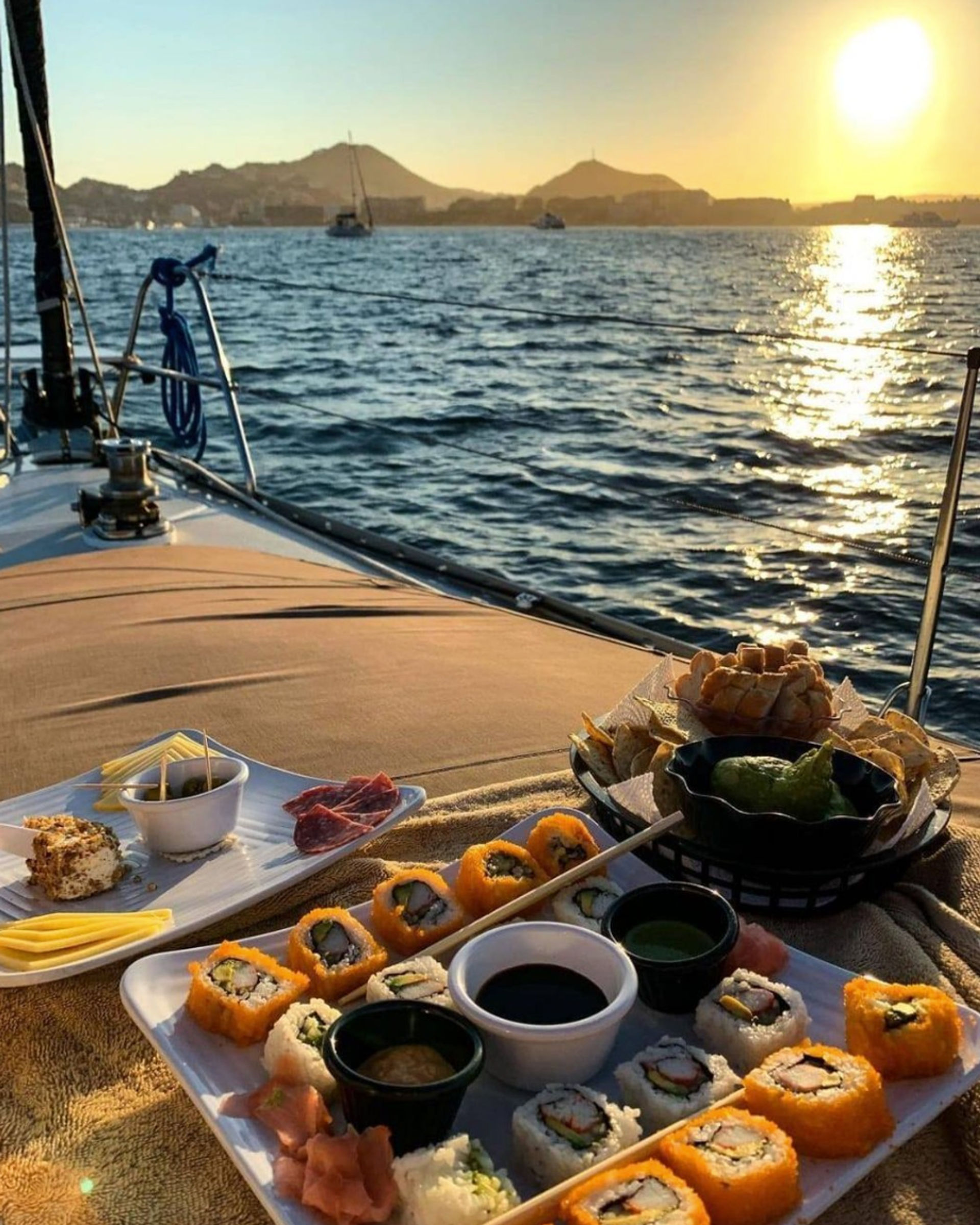 Golden-sunset sailing scene: sushi platter, dips and chips on a boat deck with sparkling ocean, anchored sailboats and hilly coastline in the distance.