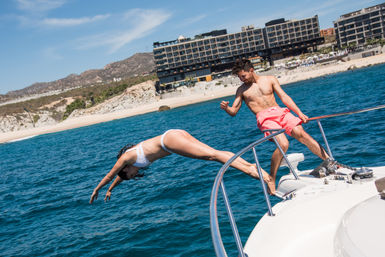 Woman in a white bikini diving off the bow of a motorboat while a man in pink swim trunks watches, turquoise ocean and rocky shoreline with a beachfront hotel under a sunny blue sky.