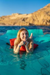 Smiling woman sun‑kissed on a turquoise floating mat in clear blue sea with sunlit rocky coastal cliffs in the background, relaxing on a red cushion under a bright sky.