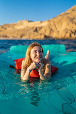 Smiling woman sun‑kissed on a turquoise floating mat in clear blue sea with sunlit rocky coastal cliffs in the background, relaxing on a red cushion under a bright sky.