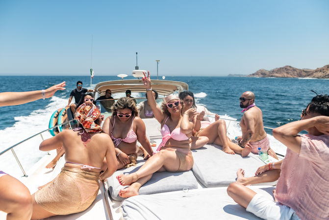 Sun-soaked yacht party on blue ocean near rocky coastline, group of friends in pink swimwear lounging and posing on the boat’s bow under a clear sky.
