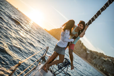 Young couple enjoying a sunset sail on the bow of a boat — man holding the wrapped mast while the woman stands barefoot on deck, sparkling ocean waves and rocky coastline behind them.