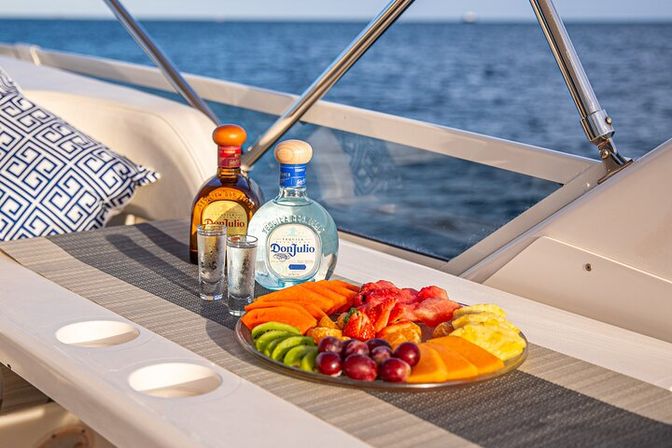 Colorful fruit platter with sliced kiwi, mango, pineapple, strawberries and grapes next to two tequila bottles and shot glasses on a yacht table with ocean view.