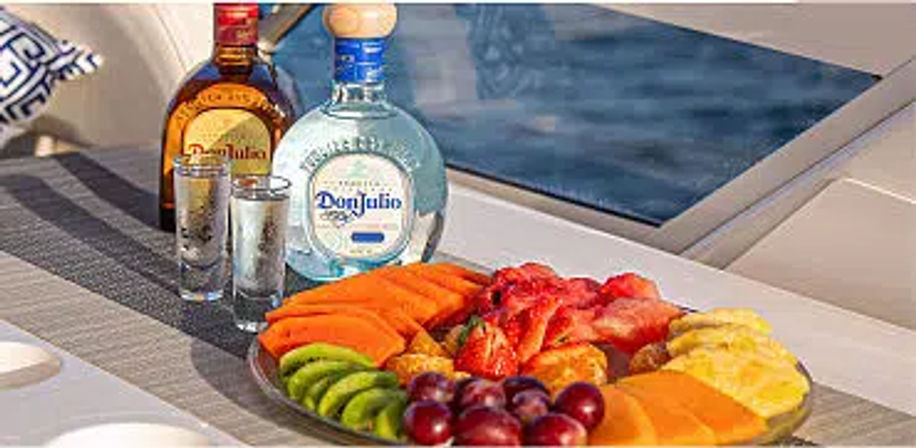 Colorful tropical fruit platter with sliced kiwi, papaya, pineapple, strawberries and grapes beside two tequila bottles and shot glasses on a yacht deck by blue water