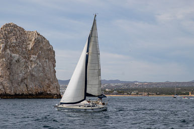 White sailboat with full sails cutting across blue ocean near towering limestone coastal cliffs, with a distant shoreline and low hills under a pale cloudy sky.