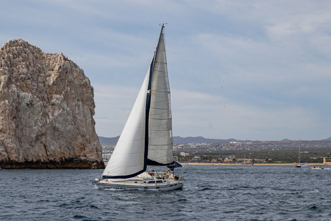 White sailboat with full sails cutting across blue ocean near towering limestone coastal cliffs, with a distant shoreline and low hills under a pale cloudy sky.