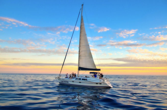 White sailboat with people enjoying a sunset cruise on a calm blue ocean, tall mainsail silhouetted against a golden-pink sky.
