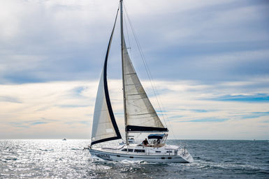 White cruising sailboat with tall mainsail and jib gliding across a sunlit open sea under a wide, partly cloudy sky.