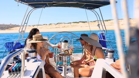 Group of friends on a sailboat enjoying drinks and snacks under wide sun hats, cruising turquoise coastal waters past a sandy shoreline on a sunny summer day