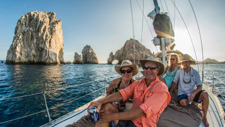 Smiling group of four relaxing on a sailboat near the sunlit rock arch and sea stacks at El Arco, Cabo San Lucas, golden-hour ocean, casual hats and drinks on deck.