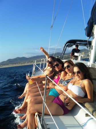 Four women in colorful swimsuits and sunglasses lounging on the rail of a sailboat, drinks in hand, legs dangling over the deep-blue ocean with a sunny sky and mountainous coastline in the background.