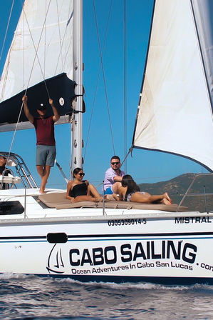 Friends lounging and sunbathing on the deck of a white sailboat with full sails on bright blue coastal waters and distant hills