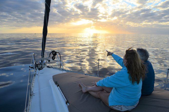 Two people relaxing on a sailboat bow at sunset, one pointing toward the golden horizon over glassy, calm ocean waters.