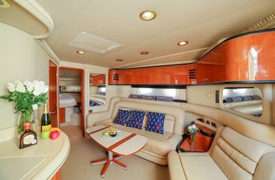 Luxury yacht interior: cozy beige leather salon with L-shaped seating, glossy wood cabinets, fold-out table with fruit and cocktail, navy anchor-pattern throw pillows, white rose bouquet and bottles on the counter, and cabin doorway in the background.
