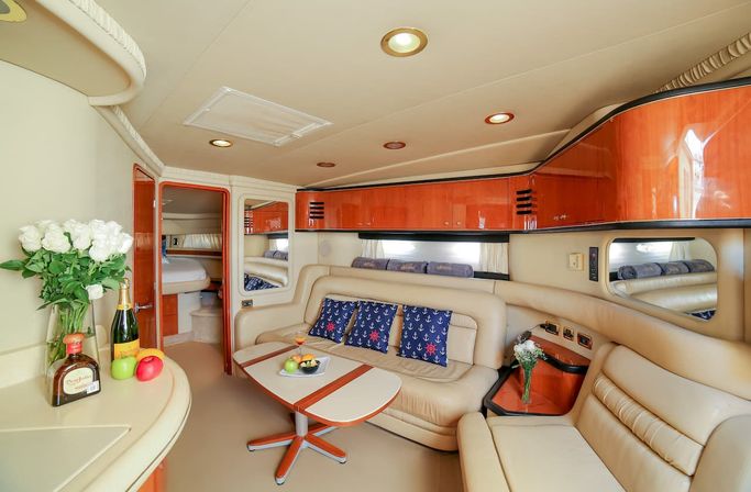 Luxury yacht interior: cozy beige leather salon with L-shaped seating, glossy wood cabinets, fold-out table with fruit and cocktail, navy anchor-pattern throw pillows, white rose bouquet and bottles on the counter, and cabin doorway in the background.