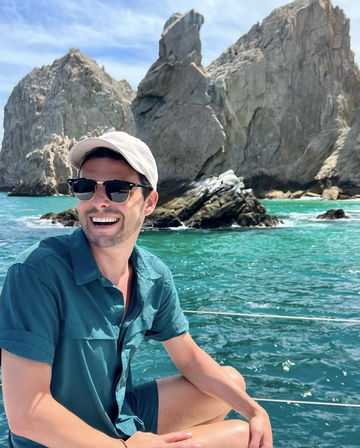 Beaming person in sunglasses and a cap on a boat, turquoise ocean and dramatic rocky sea cliffs under a sunny sky.