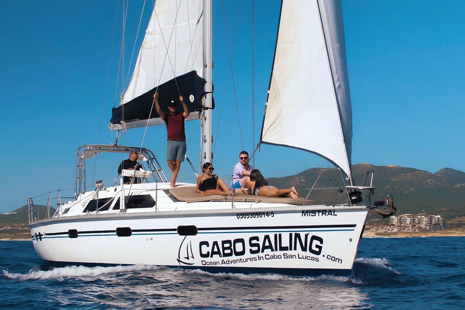 Sunlit sailboat with people relaxing on deck, sails up, cruising the ocean near Cabo San Lucas coastline