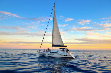 Sailing yacht with passengers cruising on calm open ocean at a colorful sunset, blue sky and pink-hued clouds reflected in the water