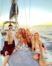 Six friends on a sailboat at sunset, lounging on the deck with drinks and a charcuterie tray as the ocean glows golden during a coastal sunset sail.