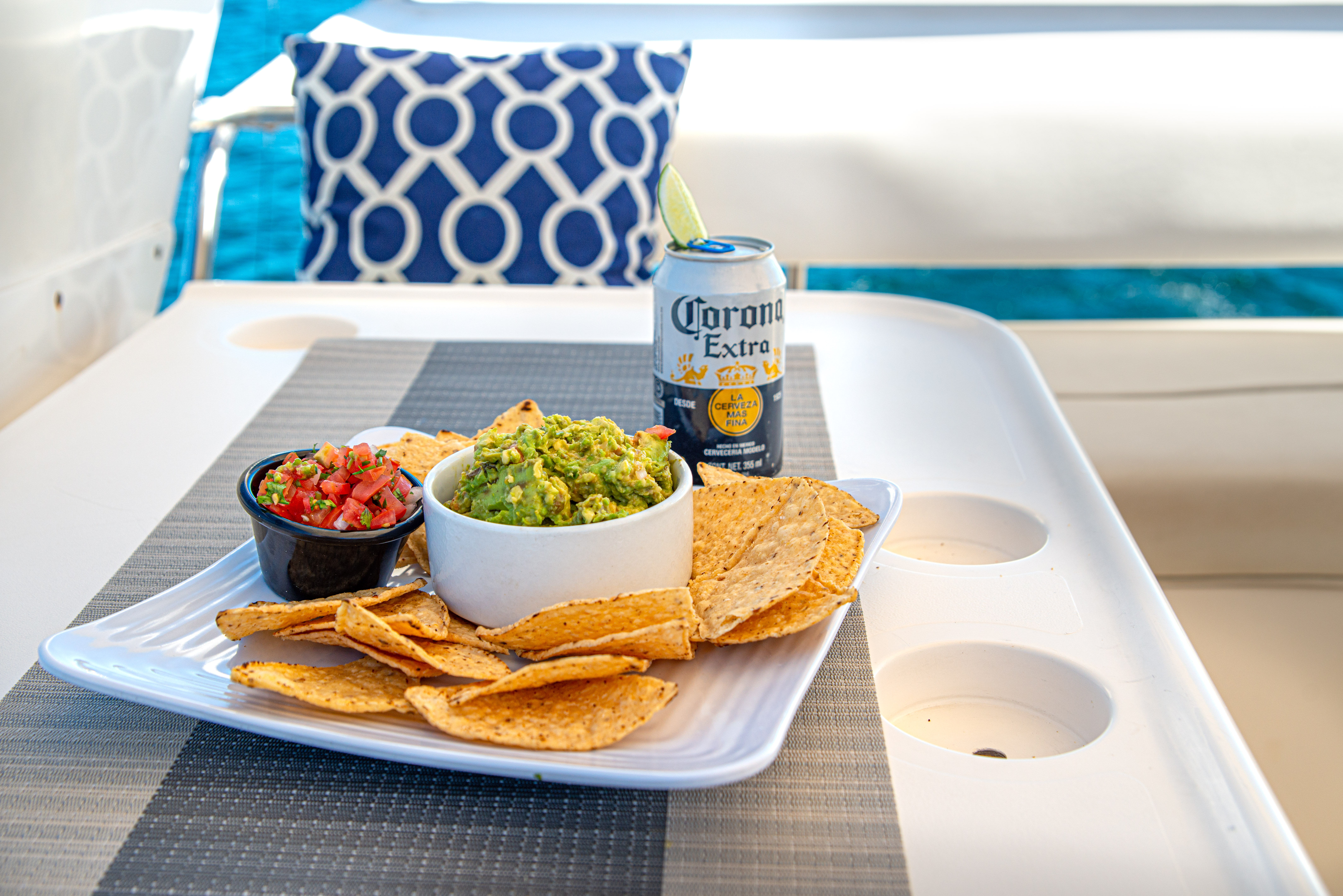 Tortilla chips with fresh guacamole and pico de gallo and a chilled beer with lime, served on a boat table with blue water and patterned cushion in the background.
