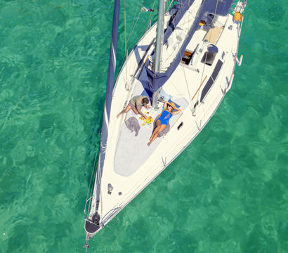Aerial view of a white sailboat on clear turquoise water with two people lounging on the deck enjoying a picnic — tropical vacation vibe.