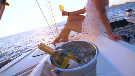 Chilled white wine in an ice bucket on a sailboat deck while a person in a white lace dress sips wine at ocean sunset, relaxing coastal cruise.