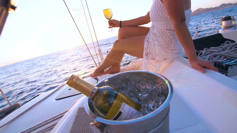 Chilled white wine in an ice bucket on a sailboat deck while a person in a white lace dress sips wine at ocean sunset, relaxing coastal cruise.