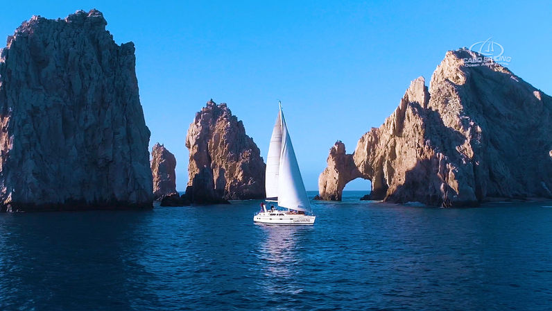 Sailboat gliding past towering sunlit sea stacks and a natural rock arch on a clear blue ocean under a bright sky