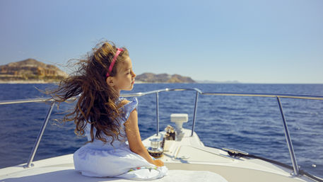 Young girl in a white sundress and pink headband sits on the bow of a yacht, hair whipping in the sea breeze as she gazes across the deep blue ocean toward a sunlit rocky coastline under a clear sky.