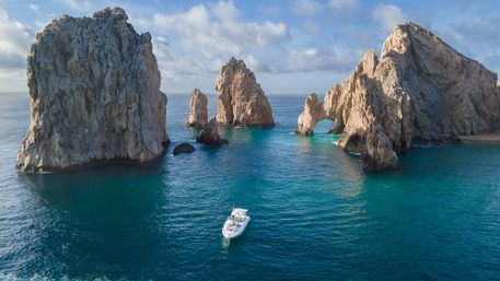 White motorboat cruising turquoise waters near towering rock arches at Land's End, Cabo San Lucas, Mexico, under a partly cloudy sky