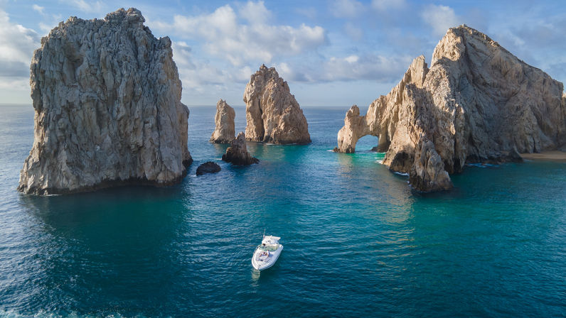 White motorboat cruising turquoise waters near towering rock arches at Land's End, Cabo San Lucas, Mexico, under a partly cloudy sky