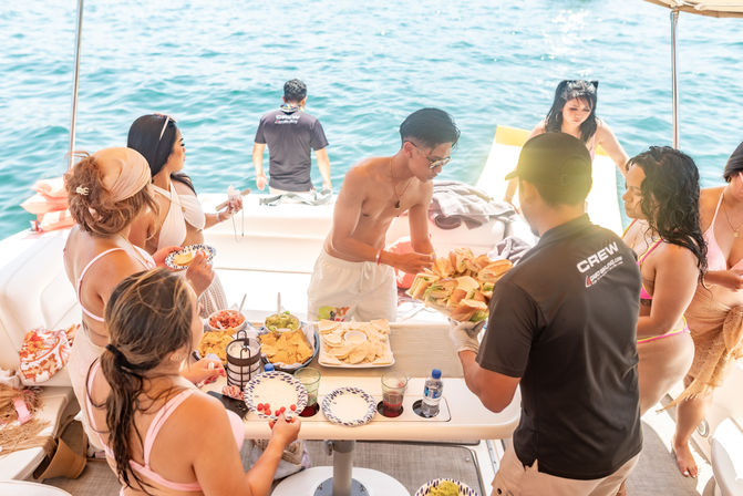 Sunny yacht party at sea with people in swimsuits gathered around a table of sandwiches, chips and dips as crew members serve food on the boat
