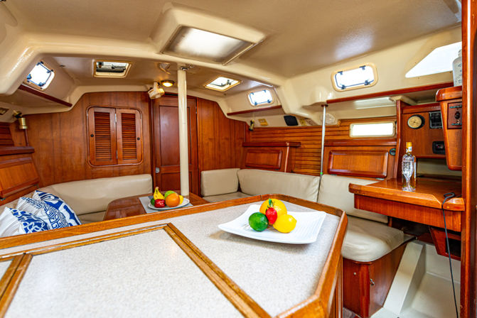 Cozy wood-paneled sailboat cabin interior with cream leather seating, center table holding a white plate of colorful fruit, porthole windows and a compact navigation station.